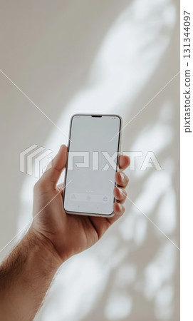 Hand holding modern smartphone with blank white screen in bright sunlight, perfect for showcasing mobile apps or social media content Hand holding modern smartphone with blank white screen in bright sunlight, perfect for showcasing mobile apps or social media content 131344097