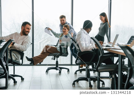 Fun at break time. Riding on a chair, smiling. Team of office workers are together indoors 131344218