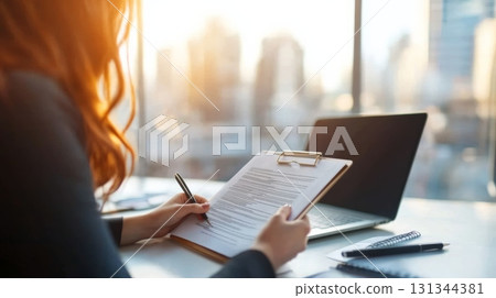 Businesswoman reviewing documents on clipboard, taking notes with pen, working at desk in modern office with laptop, sunset cityscape view through large windows 131344381