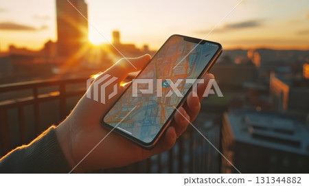 Hand holding smartphone displaying navigation app with map and location marker, overlooking cityscape at sunset from rooftop, providing sense of travel and exploration 131344882