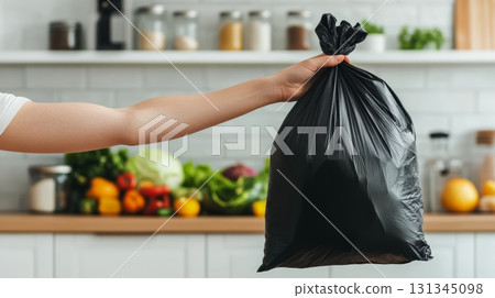 Woman holds a black plastic trash bag filled with kitchen waste, emphasizing responsible garbage disposal and waste management practices in a residential setting Woman holds a black plastic trash bag filled with kitchen waste, emphasizing responsible garbage disposal and waste management practices in a residential setting 131345098