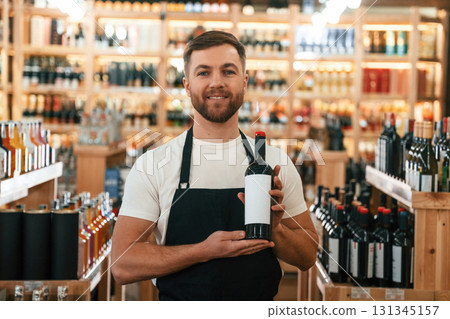 Man is holding bottle and smiling. Wine shop owner in white shirt and black apron 131345157