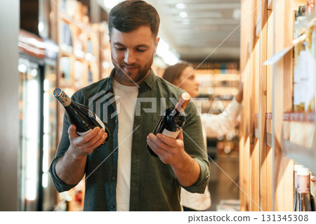 Guy holding two bottles in hands. Man and woman are choosing wine in the shop Guy holding two bottles in hands. Man and woman are choosing wine in the shop 131345308