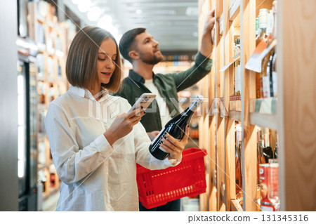 Woman is holding smartphone and bottle. Couple are choosing wine in the shop Woman is holding smartphone and bottle. Couple are choosing wine in the shop 131345316