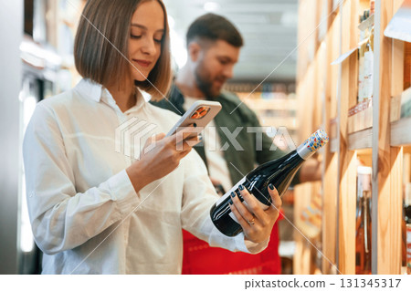 Woman is holding smartphone and bottle. Couple are choosing wine in the shop 131345317