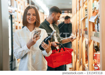 Woman is holding smartphone and bottle. Couple are choosing wine in the shop 131345318