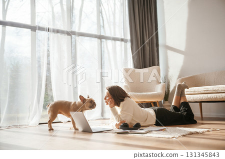 In sunlight. Lying down on floor with laptop. Looking at pet. Young woman is with her pug dog at home 131345843