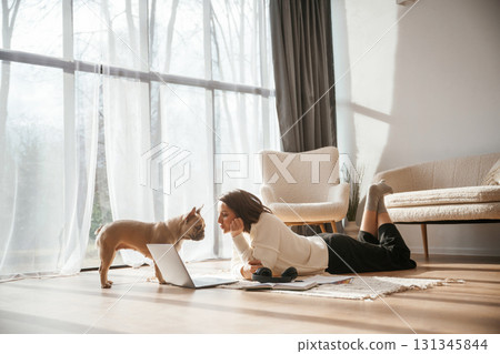In sunlight. Lying down on floor with laptop. Looking at pet. Young woman is with her pug dog at home 131345844