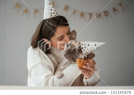 Happy owner is celebrating birthday of pet. In hat and with cake. Young woman is with her pug dog at home 131345879