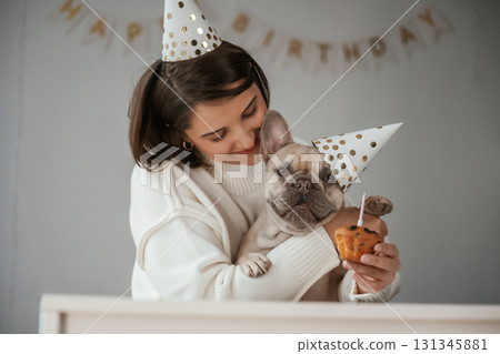 Happy owner is celebrating birthday of pet. In hat and with cake. Young woman is with her pug dog at home 131345881