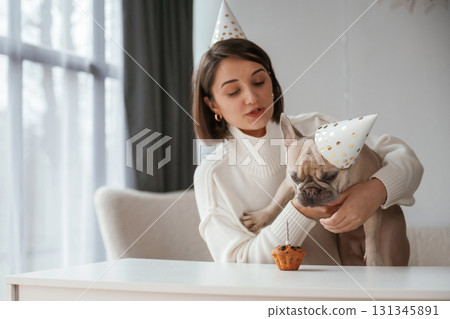 By the table, celebrating happy birthday of pet. Young woman is with her pug dog at home By the table, celebrating happy birthday of pet. Young woman is with her pug dog at home 131345891