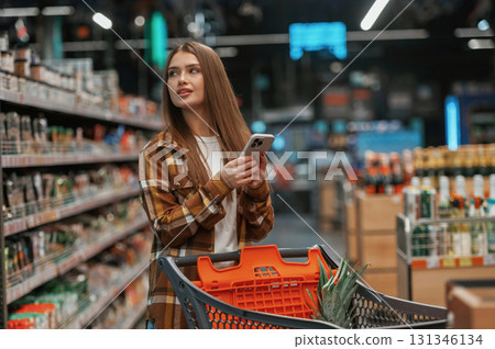 Using smartphone, standing. Woman is shopping in the supermarket Using smartphone, standing. Woman is shopping in the supermarket 131346134