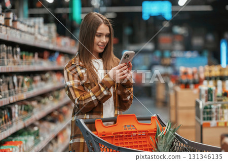 Using smartphone, standing. Woman is shopping in the supermarket Using smartphone, standing. Woman is shopping in the supermarket 131346135