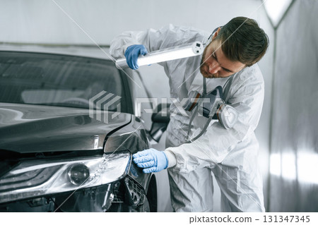 Front view. Garage worker is holding lighting and checking surface of the car Front view. Garage worker is holding lighting and checking surface of the car 131347345