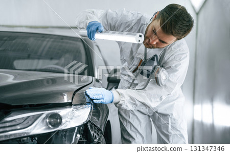 Front view. Garage worker is holding lighting and checking surface of the car Front view. Garage worker is holding lighting and checking surface of the car 131347346