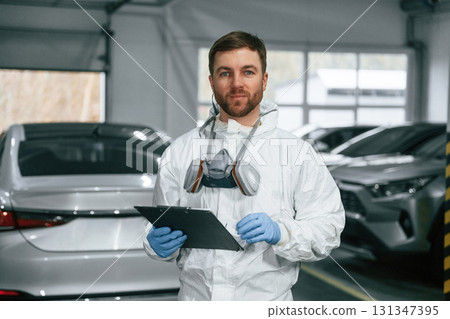 Portrait of car painter that is standing in the garage with documents in hands Portrait of car painter that is standing in the garage with documents in hands 131347395