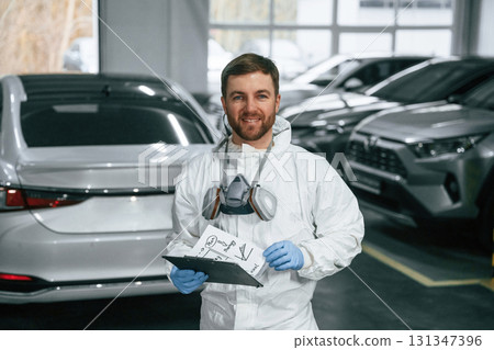 Positive facial expression. Car painter is standing in the garage with documents in hands Positive facial expression. Car painter is standing in the garage with documents in hands 131347396