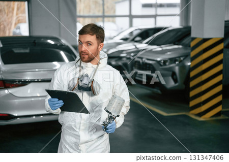 Car painter is standing in the garage with documents in hands Car painter is standing in the garage with documents in hands 131347406