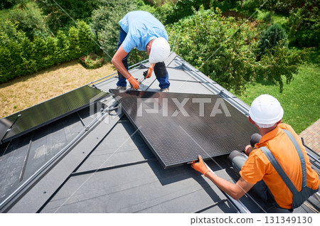 Workers building photovoltaic solar panel system on rooftop of house. Men technicians in helmets and gloves installing solar module with help of hex key outdoors. Alternative energy generation concept 131349130