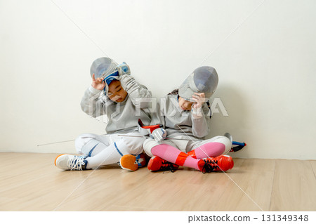 Asian boy and girl with fencing sport uniform sit beside on the floor and take mask off to relax after practice for competition. Asian boy and girl with fencing sport uniform sit beside on the floor and take mask off to relax after practice for competition. 131349348