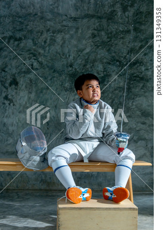 Asian boy with fencing uniform hold epee and sit on wooden bench also action of fighting to express for win in the competition. 131349358