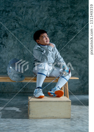 Vertical image of boy wear fencing sport uniform also hold epee and sit on wooden bench with look up to his left side. 131349359