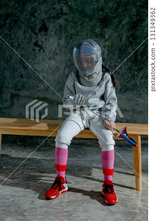 Vertical image of girl wear fencing sport uniform also hold epee and sit on wooden bench to relax after practice for the competition. 131349362