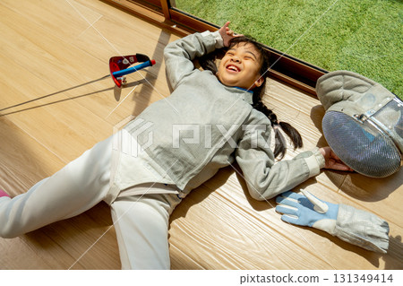 Close up of Asian girl with fencing sport uniform lie down on the floor and action like happy with smiling warm light from glass door of balcony. Close up of Asian girl with fencing sport uniform lie down on the floor and action like happy with smiling warm light from glass door of balcony. 131349414