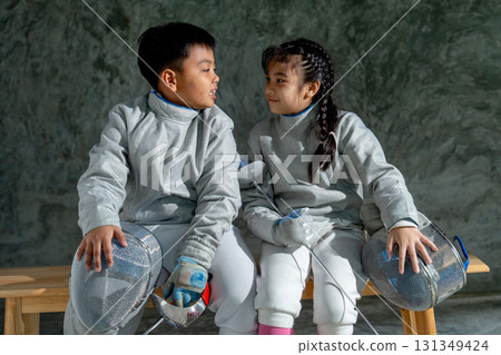 Asian brother and sister with fencing sport uniform sit beside on wooden bench and look to each other and smile also relax after practice for competition. Asian brother and sister with fencing sport uniform sit beside on wooden bench and look to each other and smile also relax after practice for competition. 131349424