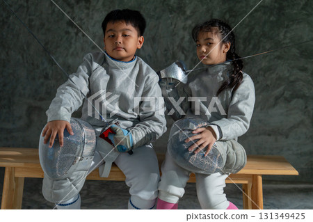 Asian brother and sister with fencing sport uniform sit beside on wooden bench and look at camera with serious emotion. Asian brother and sister with fencing sport uniform sit beside on wooden bench and look at camera with serious emotion. 131349425