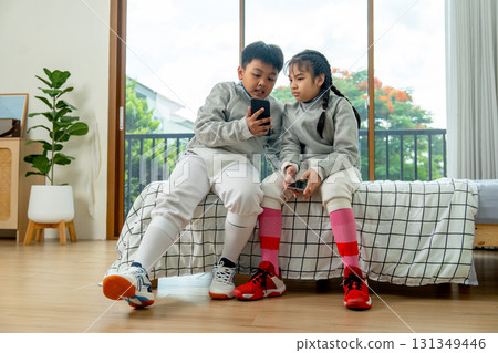 Frong view of boy and girl with uniform of fencing sport sit on bed together and discuss about the practice using mobile phone. Frong view of boy and girl with uniform of fencing sport sit on bed together and discuss about the practice using mobile phone. 131349446