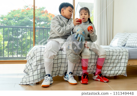 Boy and girl with uniform of fencing sport sit on bed together during enjoy eating apple in bedroom during relax after practice. 131349448