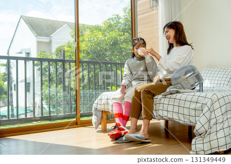 Asian mother sit on bed next to her daughter and give a glass of mild to her child during practice fencing sport in bedroom with soft morning light and happiness. 131349449