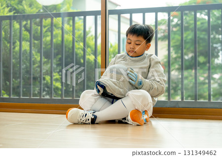 Side view of Asian boy sit on the floor in front of glass door near balcony of his house also hold helmet of fencing sport with soft light. 131349462
