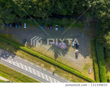 Aerial view of parking lot beside forest, with aligned vehicles, central triangular monument, hedges, and road markings blending geometry and natural surroundings. Aerial view of parking lot beside forest, with aligned vehicles, central triangular monument, hedges, and road markings blending geometry and natural surroundings. 131349633