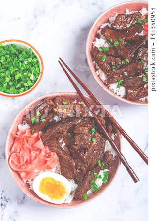Gyudon Japanese beef with rice, pickled ginger, boiled egg slice, garnished with green onions, in a bowl with chopsticks, 131350953