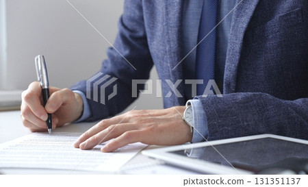 Professional businessman signing contract with silver pen, wearing tailored suit, business documents and digital tablet nearby. Business people concept Professional businessman signing contract with silver pen, wearing tailored suit, business documents and digital tablet nearby. Business people concept 131351137