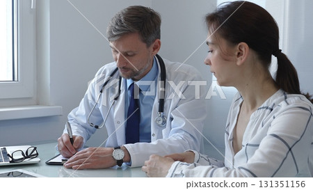 Male doctor writing notes on a clipboard while attentively listening to a female patient during a medical consultation in a hospital office, fostering communication and understanding 131351156