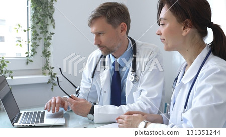 Two doctors are studying medical charts on a laptop, discussing patient data in a hospital or clinical setting. Medicine and healthcare concept. 131351244