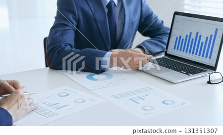 Business colleagues or partners are discussing financial data on a laptop and printed documents while analyzing charts and market trends at office table. Businesspeople and finance concept 131351307