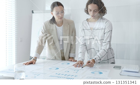 Professional businesswomen analyzing financial charts, collaborating in sleek corporate workspace, highlighting data. Business people concept 131351341