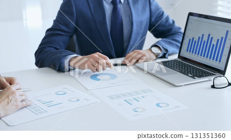 Business colleagues or partners are discussing financial data on a laptop and printed documents while analyzing charts and market trends at office table. Businesspeople and finance concept Business colleagues or partners are discussing financial data on a laptop and printed documents while analyzing charts and market trends at office table. Businesspeople and finance concept 131351360