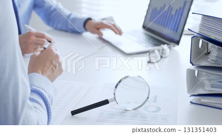 Business team working together, analyzing financial data on charts and documents, laptop and folders on white desk in office, close up of magnifying glass. Audit and taxes theme in business Business team working together, analyzing financial data on charts and documents, laptop and folders on white desk in office, close up of magnifying glass. Audit and taxes theme in business 131351403