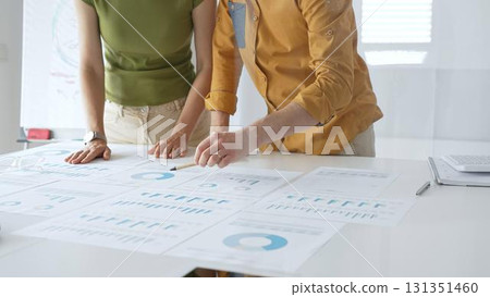 Business professionals analyzing financial reports, pointing at data points on charts and graphs lying across white desk surface 131351460