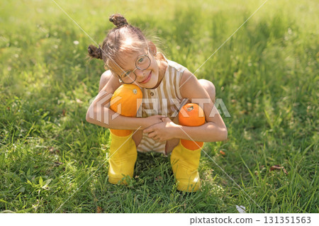 A cute, cheerful girl in rubber boots hugs two large yellow squash. The harvest season in the vegetable garden A cute, cheerful girl in rubber boots hugs two large yellow squash. The harvest season in the vegetable garden 131351563