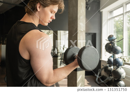 Muscular young caucasian man lifting dumbbell in gym setting. Weightlifting exercises, physical fitness. Work out. 131351750
