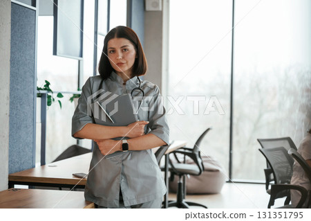 Standing and holding notepad. Female doctor in coat is indoors Standing and holding notepad. Female doctor in coat is indoors 131351795