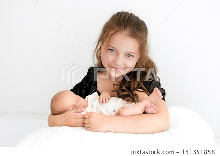 A cute beautiful girl holds her newborn baby brother in arms while sitting in bed. Love and help in caring for younger children A cute beautiful girl holds her newborn baby brother in arms while sitting in bed. Love and help in caring for younger children 131351858