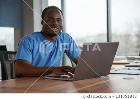 Looking at the camera and smiling. Doctor is sitting by the table with laptop in clinic 131351918