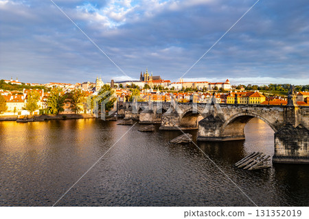 Early morning light casts a glow on Prague Castle and Charles Bridge, reflecting beauty over the Vltava River. The old town awakens as clouds drift above the historic skyline. 131352019